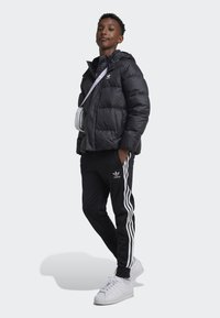 Black padded jacket with hood, white logo, black pants with white side stripes, and white sneakers. Simple design with a relaxed fit.