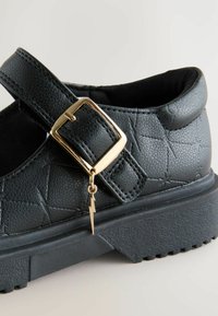 Chaussure en simili cuir noir avec une semelle épaisse, ornée d'une sangle à boucle dorée et d'un charm en forme d'éclair. Surface texturée avec des motifs géométriques.
