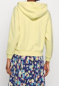 Gul hooded sweatshirt med långa ärmar, strukturerad tyg och ribbade muddarna. Kombinerad med en blommig veckad kjol i blått och flera färger.