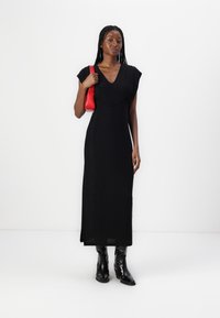 Black maxi dress with short sleeves, V-neck, and draped waist. Model holds a red handbag. Black ankle boots complete the look.