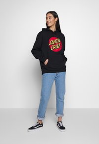 Agasalho preto com um logotipo circular vermelho e amarelo "SANTA CRUZ". Combinado com jeans azuis com as mangas enroladas e sapatilhas pretas.