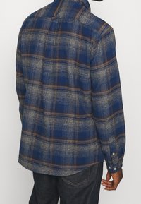Blå och grå rutigt flanellskjorta med långa ärmar, knäppning vid ärmsluten och rundad nederkant. Har bruna inslag i rutmönstret.