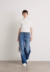 Hvit kortermet høyhalset genser og lyseblå vide jeans. Modellen holder en lys veske i den ene hånden og har på seg hvite sneakers. Enkelt bakgrunn.