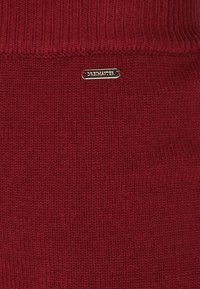 Țesătură tricotată burgundy cu o textură ribbed. Prezintă un mic etichetă din metal auriu cu inscripția "DREAMMASTER". Design simplu și solid.
