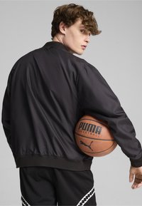 Sort bomberjakke med en glat tekstur, ribbede manchetter og kant. Modellen holder en basketball. Atletiske shorts med et mønstrede sidelayout.