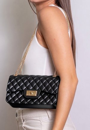 Vrouw draagt een witte mouwloze top en lichte spijkerbroek, met een zwarte doorgestikte leren schoudertas met gouden kettingriem en sluiting.