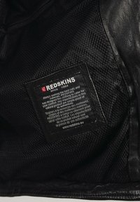 Intérieur d'une veste en cuir noir montrant une doublure en maille et une étiquette rectangulaire avec du texte blanc et rouge, détaillant la marque et les instructions d'entretien.