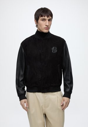 Homme aux cheveux bruns courts portant une veste universitaire noire à manches en cuir et un logo "B", assortie à un pantalon beige, debout sur fond blanc.