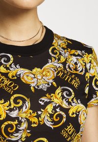 Czarna koszula krótki rękaw z wzorem barokowym w kolorze złotym i białym oraz nadrukowanym tekstem "Versace Jeans Couture" w pogrubionej czcionce.