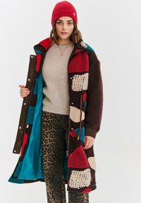 Žena v pestrofarebnom patchwork fleece kabáte, béžovom svetri, nohaviciach s leopardím vzorom a červenom pletenom čiapke, stojaca na bielom pozadí.
