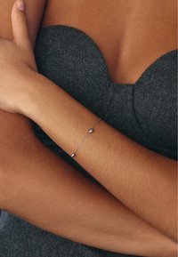 Silver chain bracelet with two small heart-shaped accents, worn on a tanned arm, against a textured gray background.