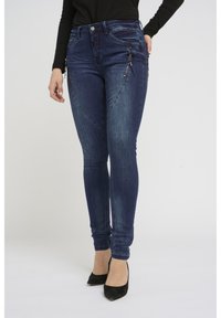 Jean skinny en denim bleu foncé avec des zips sur les côtés, une taille haute et un léger délavage. Assortis à des talons noirs. Texture lisse dans l'ensemble.