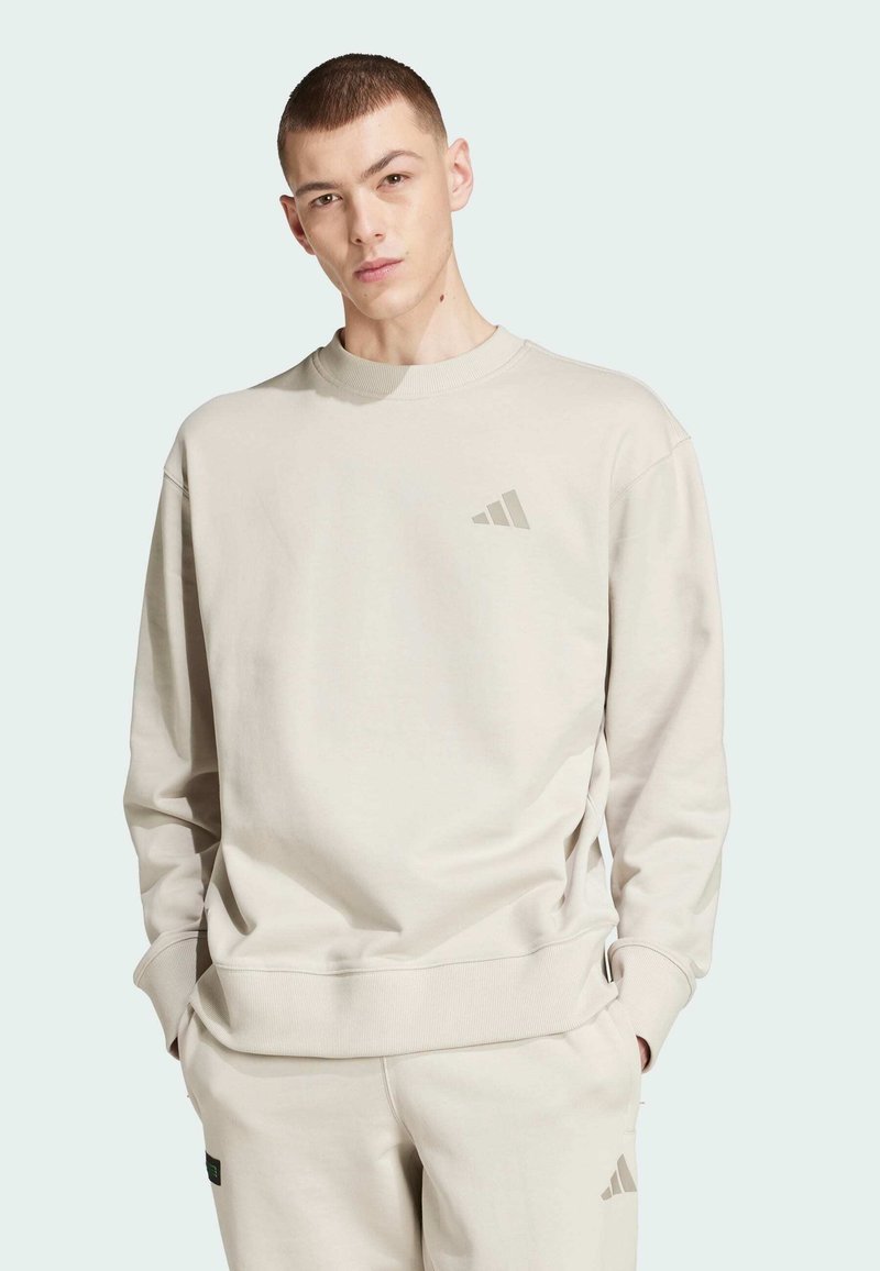 Sudadera beige claro hecha de tejido suave, con cuello redondo, mangas con puños y un sutil logo en el pecho. Diseño sencillo en toda la prenda.