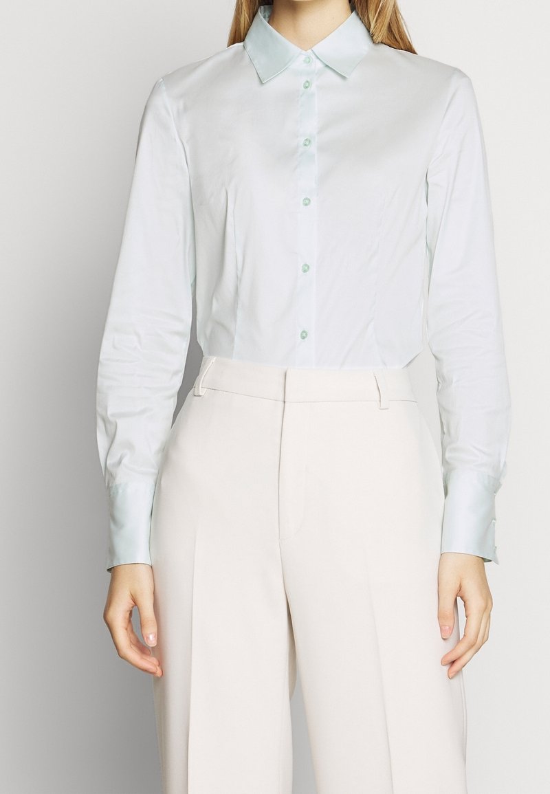 Chemise bleu clair à manches longues avec un col, devant boutonnière et texture lisse. Associée à un pantalon beige taille haute.