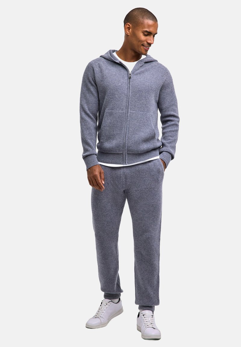 Grau gestrickter Kapuzen-Sweatshirt mit Reißverschluss und Fronttaschen, kombiniert mit passenden, schmal zulaufenden Hosen. Dazu weiße Sneaker und ein weißes T-Shirt mit Rundhalsausschnitt darunter getragen.