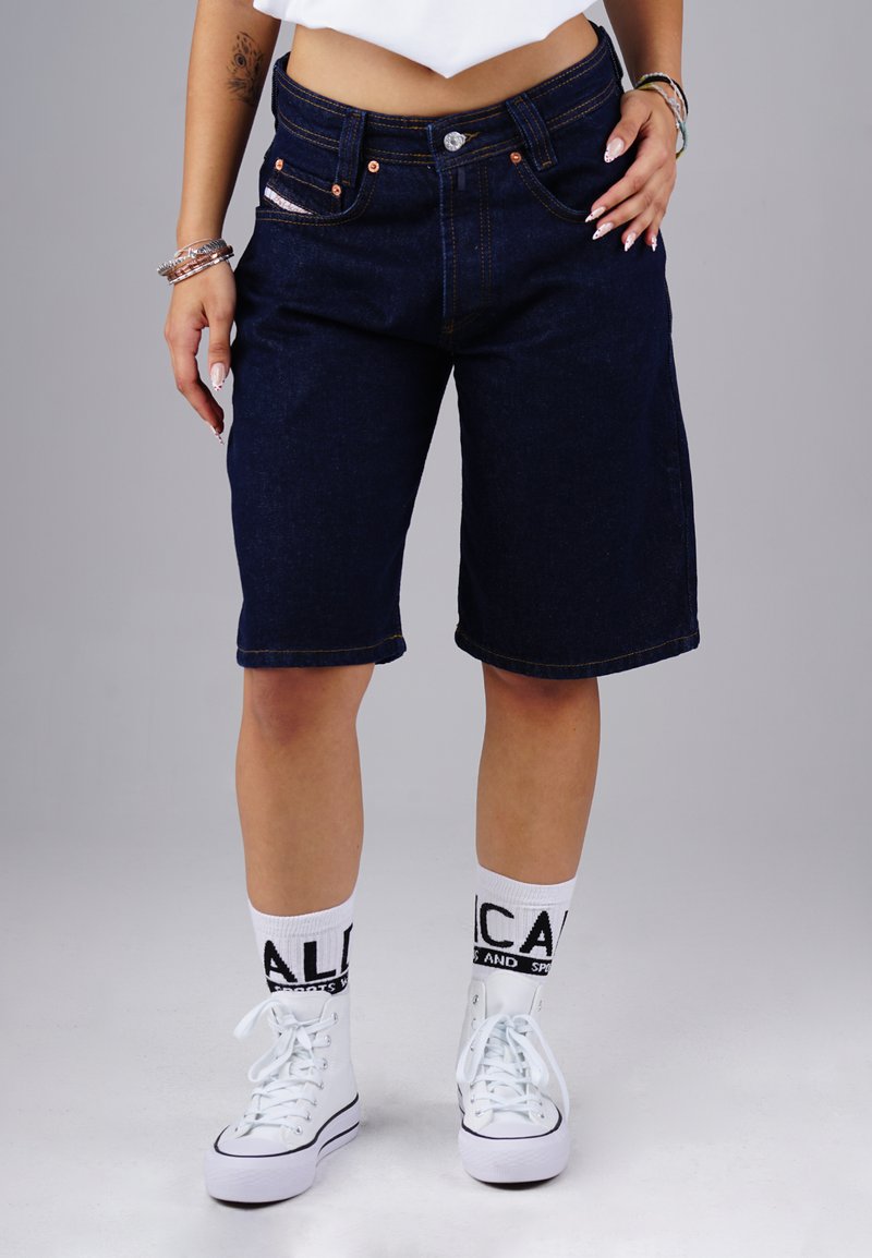 Person wearing dark denim knee-length shorts, white printed socks, and white high-top sneakers standing against a plain background.