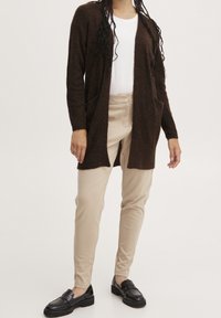 Cárdigan de punto marrón con abertura en la parte delantera y dos bolsillos frontales, combinado con pantalones beis y mocasines negros, presentando texturas suaves y un ajuste casual.