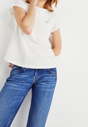 Person iført en hvid t-shirt med korte ærmer og blå cowboybukser, poser med den ene arm bøjet og den anden bag ryggen.