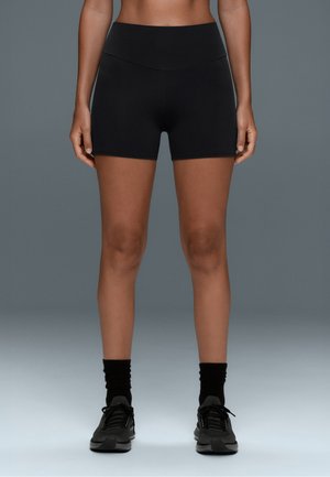 Femme debout portant un short de sport noir taille haute, des chaussettes noires et des baskets noires sur un fond gris.