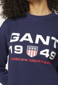 Námořnická modrá mikina s velkým bílým textem "GANT," "1949" a "AMERICAN SPORTSWEAR," se štítem v červené a modré barvě.