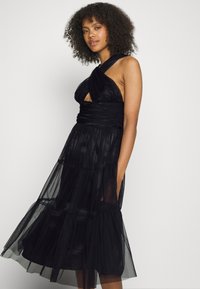 Vestido preto de alças com detalhes recortados, tecido de tule em camadas, comprimento até o meio da perna, textura suave e um corpete justo que transita para uma saia esvoaçante.