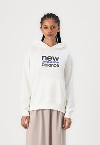 Baltas kapucis džemperis ar priekšējo kabatu, ar izteiksmīgu melnu un zilu "New Balance" logo. Gluda tekstūra un brīva piegriezuma dizains.