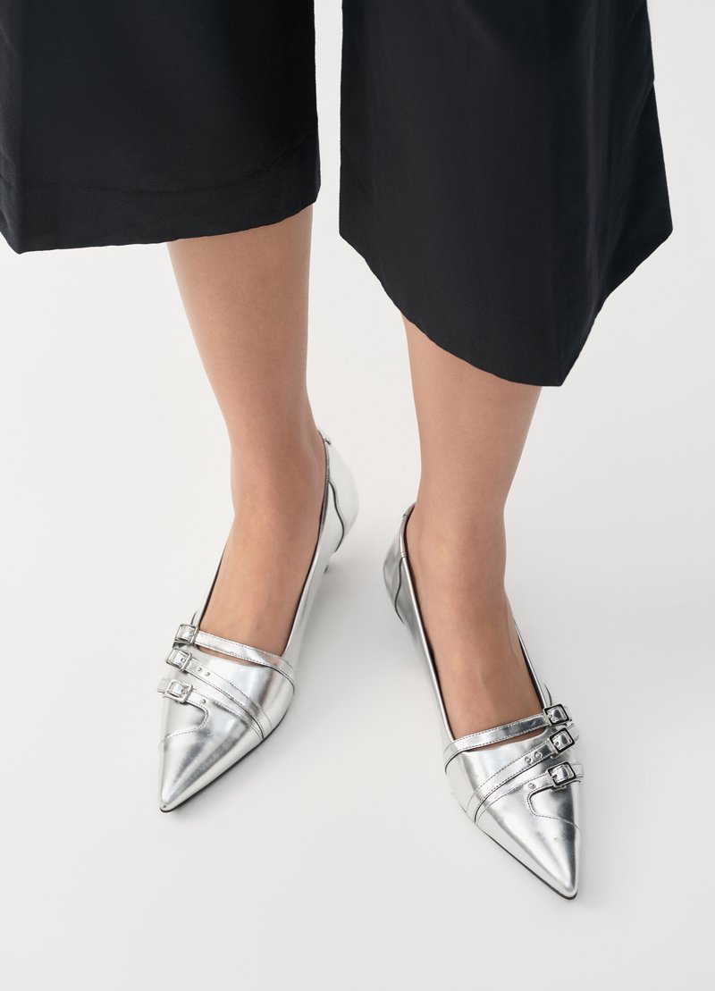 Ballerine metalliche argento con punte appuntite, dotate di due cinghie regolabili con fibbie, texture liscia e tacco basso. Visibili pantaloni neri.