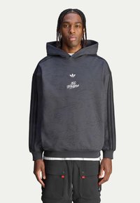 Mustassa hupparissa on sävyttävä maastokuvio, brodeerattu logo rinnassa ja kolme raidallista yksityiskohtaa hihoissa.