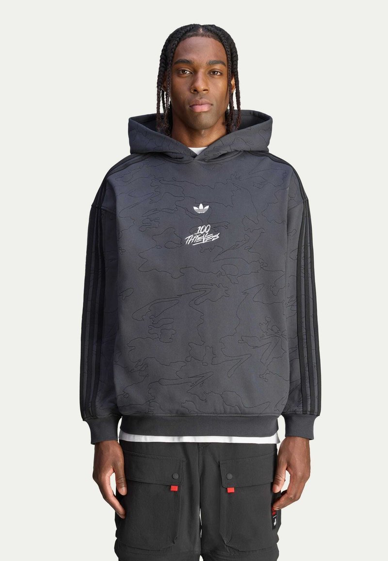 Mustassa hupparissa on sävyttävä maastokuvio, brodeerattu logo rinnassa ja kolme raidallista yksityiskohtaa hihoissa.