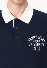 Sötétkék polo ing fehér gallérral, három gombbal, és fehér betűkkel hímzett "TOMMY JEANS UNIVERSITY CLUB" felirattal. Texturált anyag.