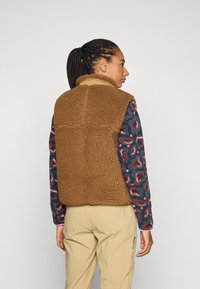 Gilet en polaire marron avec un col haut, présentant un tissu texturé et une coupe décontractée, porté par-dessus une chemise à manches longues à motifs et un pantalon clair.