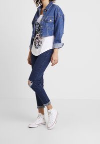 Chaqueta de denim, diseño corto, azul claro; camiseta blanca con estampado floral; jeans skinny azul oscuro con rodillas desgastadas; zapatillas altas blancas.