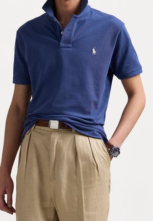 Persoon die een blauw poloshirt met een logo draagt, beige geplooide broek, een bruine riem en een polshorloge aan de linkerpols.