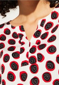 Blusa blanca con un patrón de remolinos en rojo y negro, con un escote en V y una tapeta detallada. La tela parece suave y ligera.