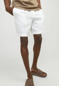 Shorts en lin blancs avec une coupe décontractée et des poches latérales, présentant un design simple et finis avec des ourlets. Assortis avec des sandales brunes.