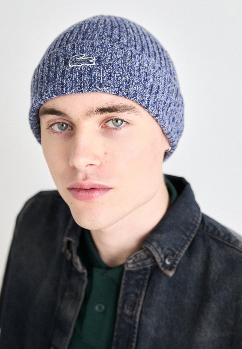 Bonnet en tricot dans des tons de bleu, avec une texture côtelée et un petit logo brodé. Porté avec une veste en denim foncé sur une chemise verte.