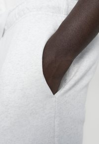 Svetlosivá textúrovaná látka s viditeľnou vreckom a tmavohnedou pažou vyčnievajúcou zo strany. Jednoduchý, neformálny dizajn.