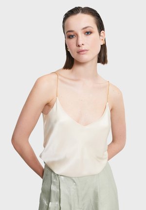 Młoda kobieta z krótkimi, mokrymi włosami, ubrana w kremową satynową koszulkę na cienkich złotych łańcuszkach oraz jasne, zielone błyszczące spodnie, z rękami za plecami.