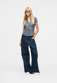 Femme aux longs cheveux blonds et portant des lunettes, habillée d'un t-shirt gris ajusté à manches courtes, d'un jean large bleu foncé et de baskets blanches.