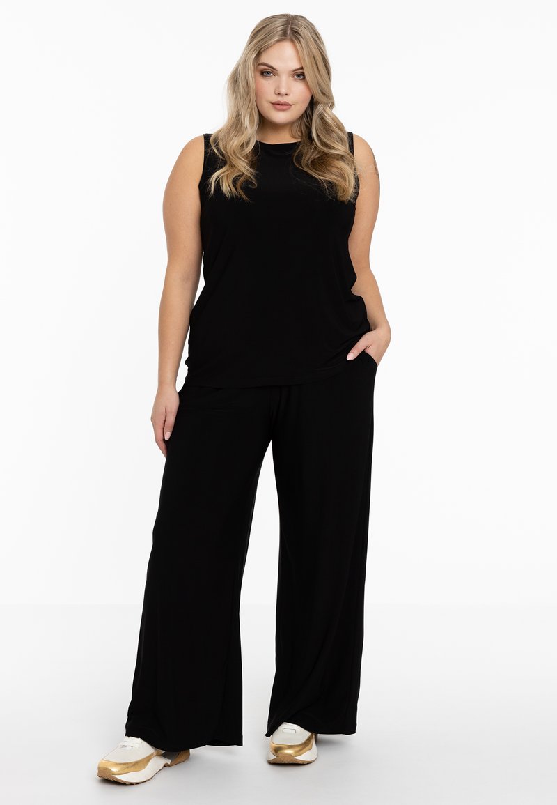 Zwarte mouwloze top gecombineerd met losse zwarte broek, beide gemaakt van zachte stof. Schoenen zijn wit met gouden accenten.