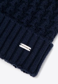 Pletemá tmavě modrá čepice s texturovaným vzorem a žebrovaným lemem; na boku je stříbrná štítek s logem "WITTCHEN".