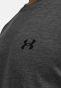 Harmaa urheilu paita, joka on valmistettu teksturoidusta kankaasta ja jossa on musta Under Armour -logo rinnassa. Lyhyet hihat, pyöreä kaula-aukko.