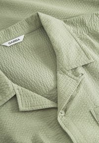 Camicia verde testurizzata con colletto, dettagli con bottoni e motivo sottile. L'etichetta riporta "GABBA". Il tessuto sembra leggero e intrecciato.