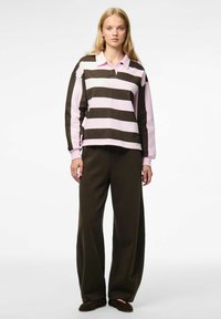 Femme blonde se tient debout, portant un polo à rayures larges rose et marron foncé à manches longues, avec un pantalon large marron foncé et des chaussures sombres.