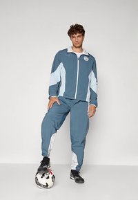 Blå och ljusblå träningsoverall med zipjacka och matchande byxor med abstrakta vita mönster på sidorna. Svart fotboll nedanför.