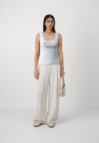 Top sin mangas blanco acanalado con un logo plateado de "Guess", combinado con pantalones sueltos de color crema y zapatos ligeros con textura.