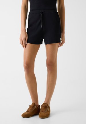 Vrouw die zwarte shorts met trekkoord en bruine suède sneakers draagt, staand tegen een effen lichte achtergrond.