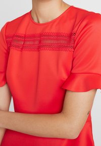 Robe rouge en tissu lisse, avec un détail en dentelle sur la poitrine et de courtes manches volantes.
