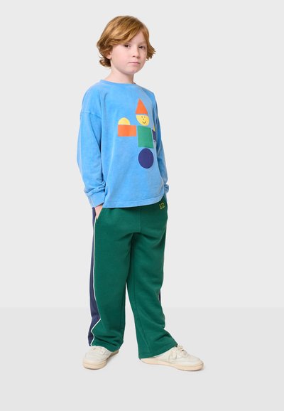 Plava košulja dugih rukava s šarenim geometrijskim oblicima i smiješkom, uparen sa zelenim trenirkama s tamnoplavim bočnim prugama i džepom.