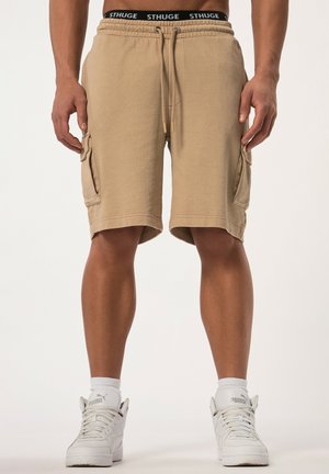 Shorts cargo en khaki avec une taille à cordon, des poches latérales et une texture en coton lisse. Associés à des baskets blanches et des chaussettes mi-mollet.